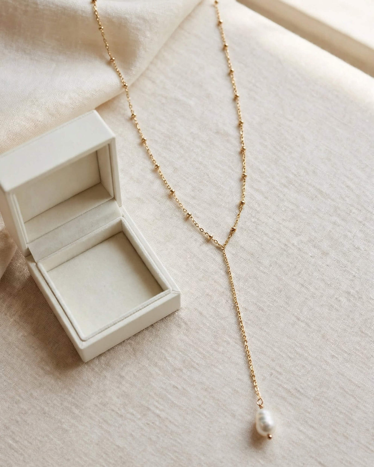 A delicate gold Y-necklace with a single baroque pearl pendant, displayed next to an open cream-colored jewelry box on a soft beige fabric background.
