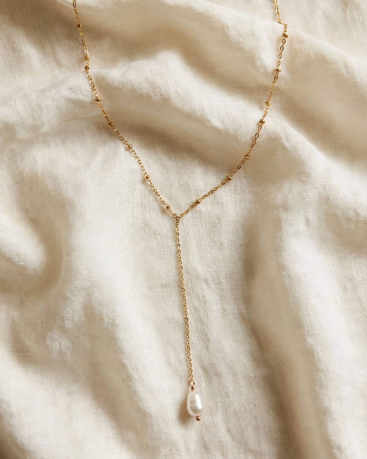 A delicate gold Y-necklace featuring a single baroque pearl pendant, laid flat on a textured cream linen fabric background.