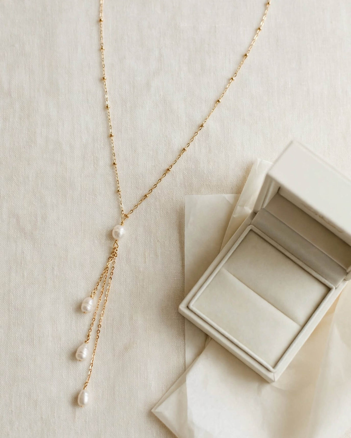 A delicate gold Y-necklace with three dangling pearl strands, displayed next to an open cream-colored jewelry box on a textured beige fabric background.