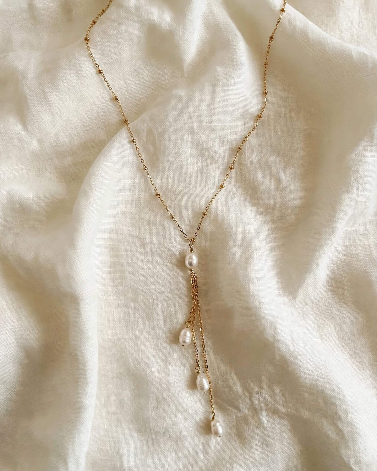 A delicate gold chain necklace with a central pearl and three cascading pearl drops, laid flat on a textured off-white linen fabric background.