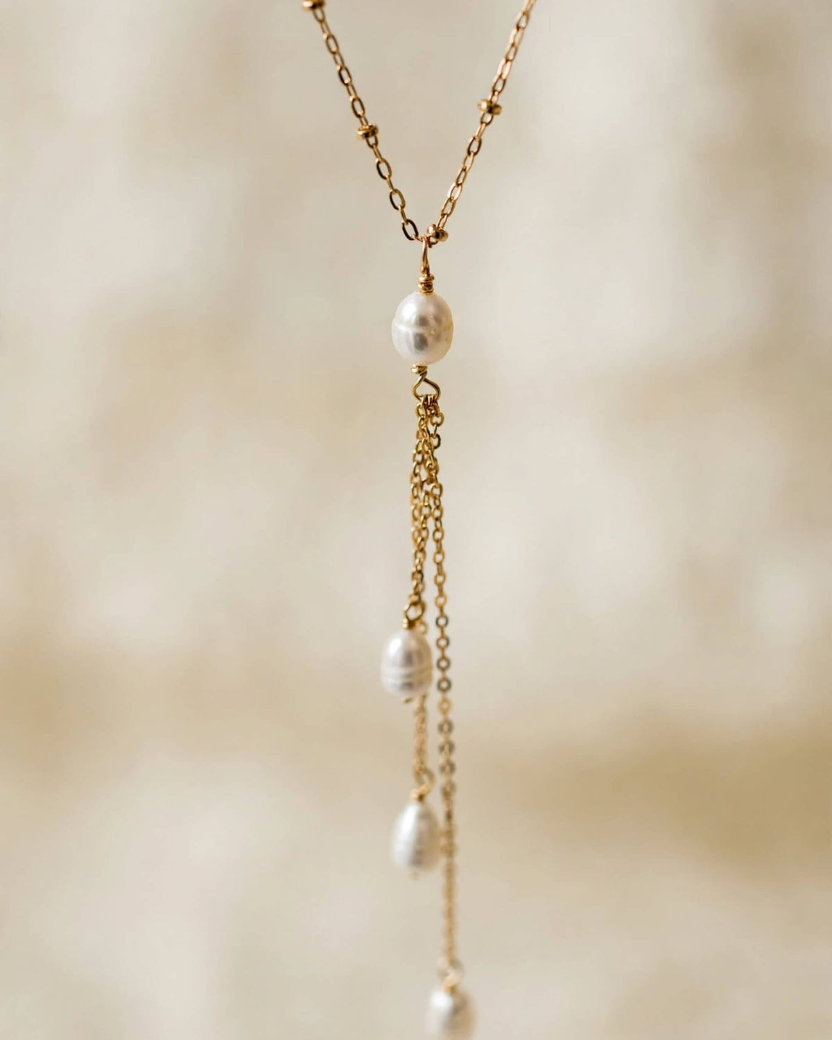 A gold chain necklace featuring a central white pearl and three cascading dangle chains with small white pearls, set against a soft, warm beige background.