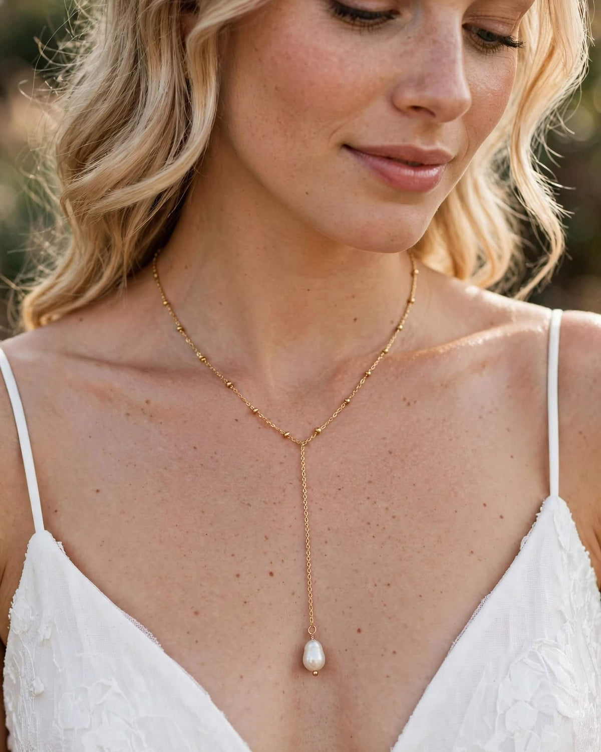 A close-up lifestyle shot of a woman wearing a delicate gold lariat necklace with a single white freshwater pearl pendant, styled with a white lace camisole.