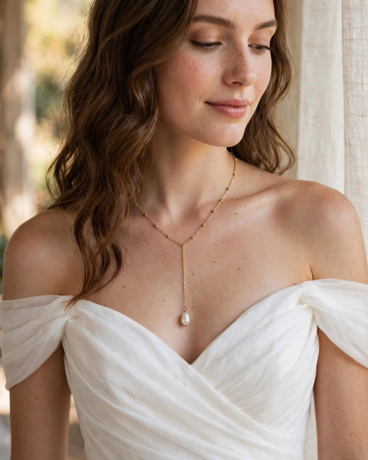 A woman wearing a delicate gold lariat necklace with a single white pearl pendant, styled with an off-the-shoulder white dress.