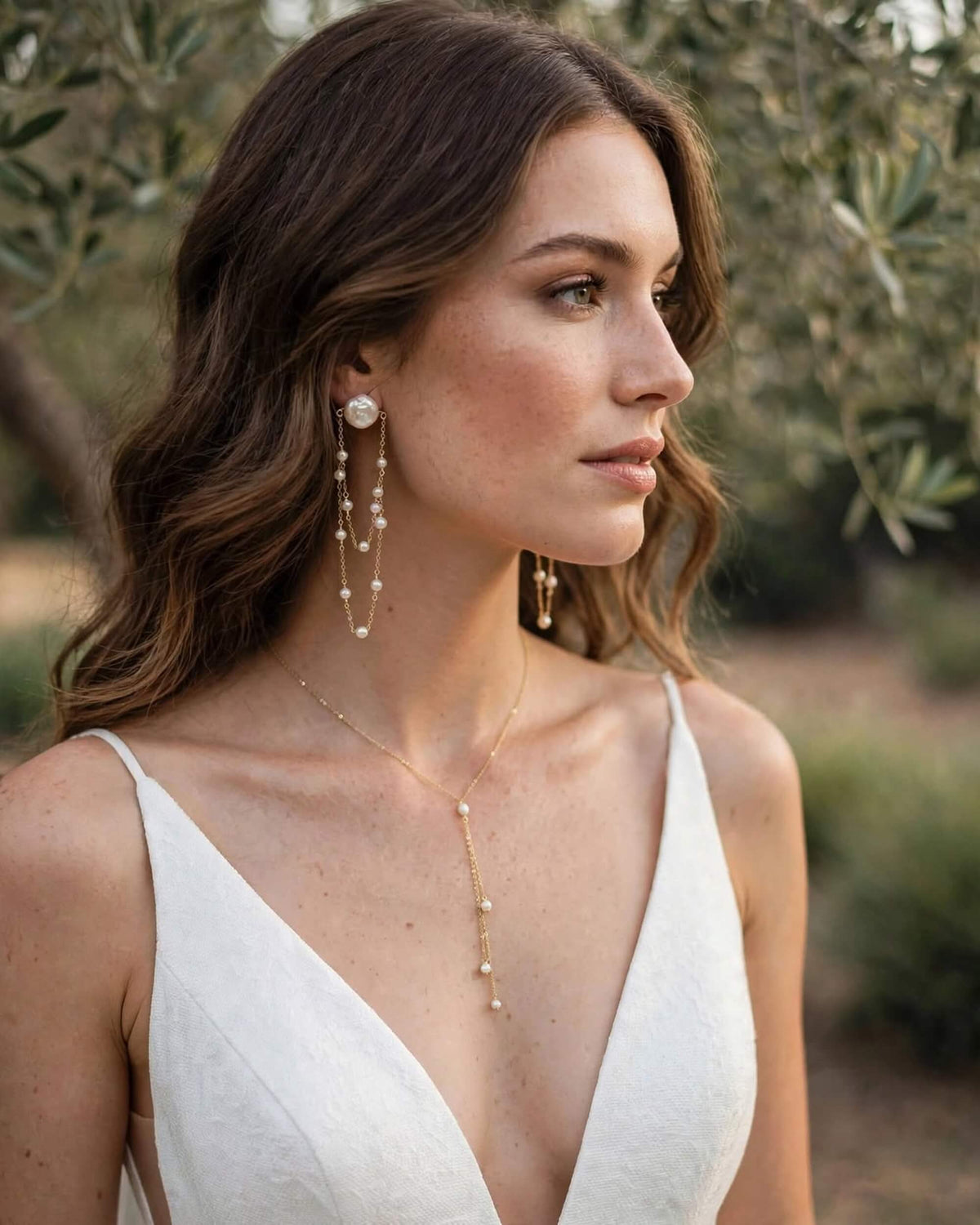 A woman in a white wedding dress wearing elegant gold jewelry, including a delicate gold lariat necklace and long pearl drop earrings, set against a soft-focus olive garden background.