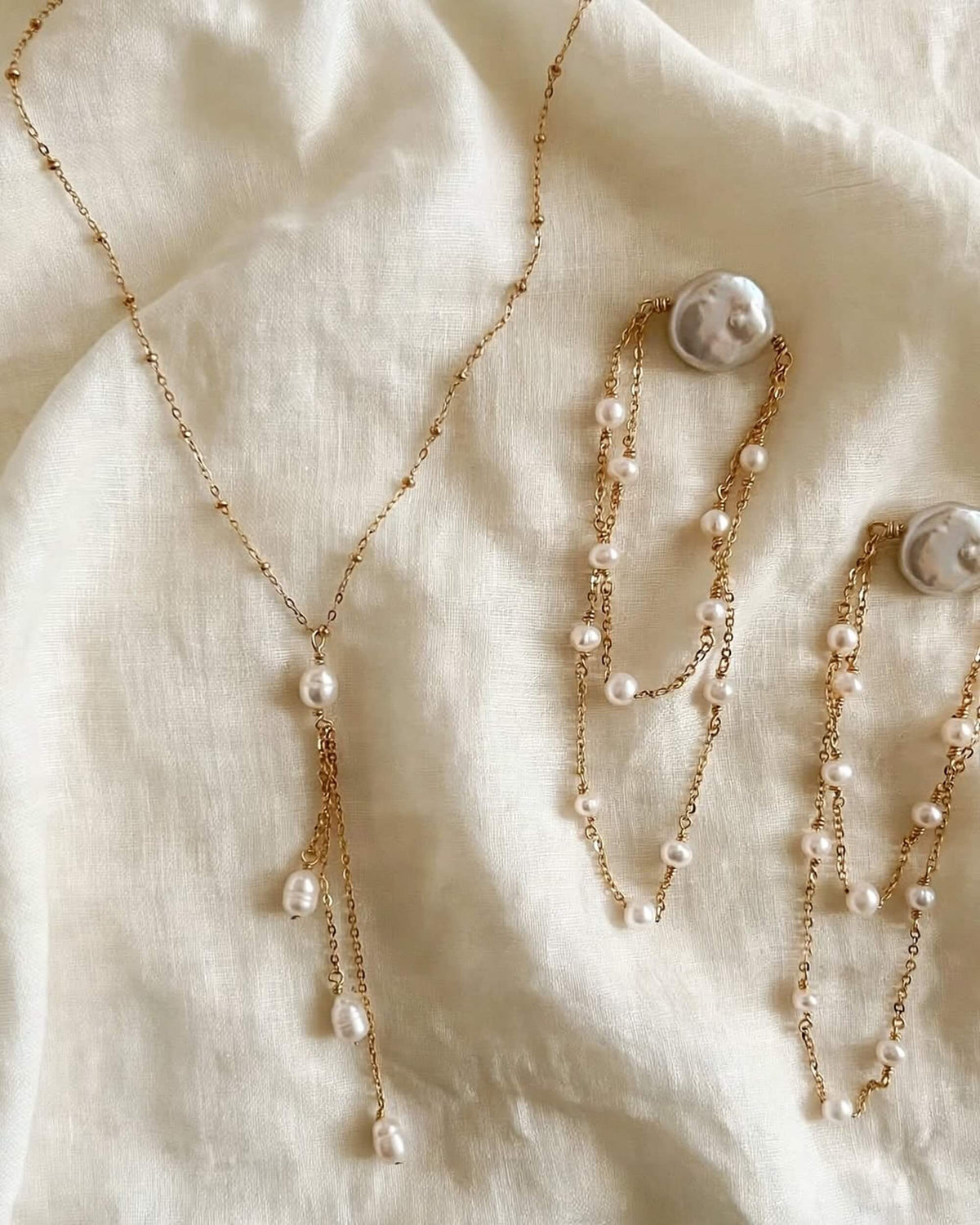 A gold-tone jewelry set featuring a lariat-style necklace and matching earrings with irregular white freshwater pearls, displayed on a draped cream linen fabric background.