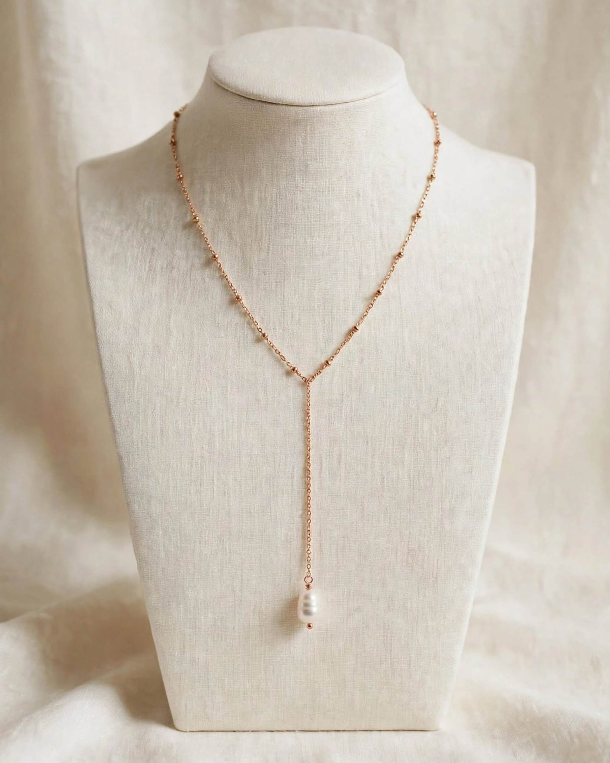 A delicate rose gold Y-necklace with a single white pearl pendant, displayed on a cream fabric-covered jewelry bust against a soft beige draped background.