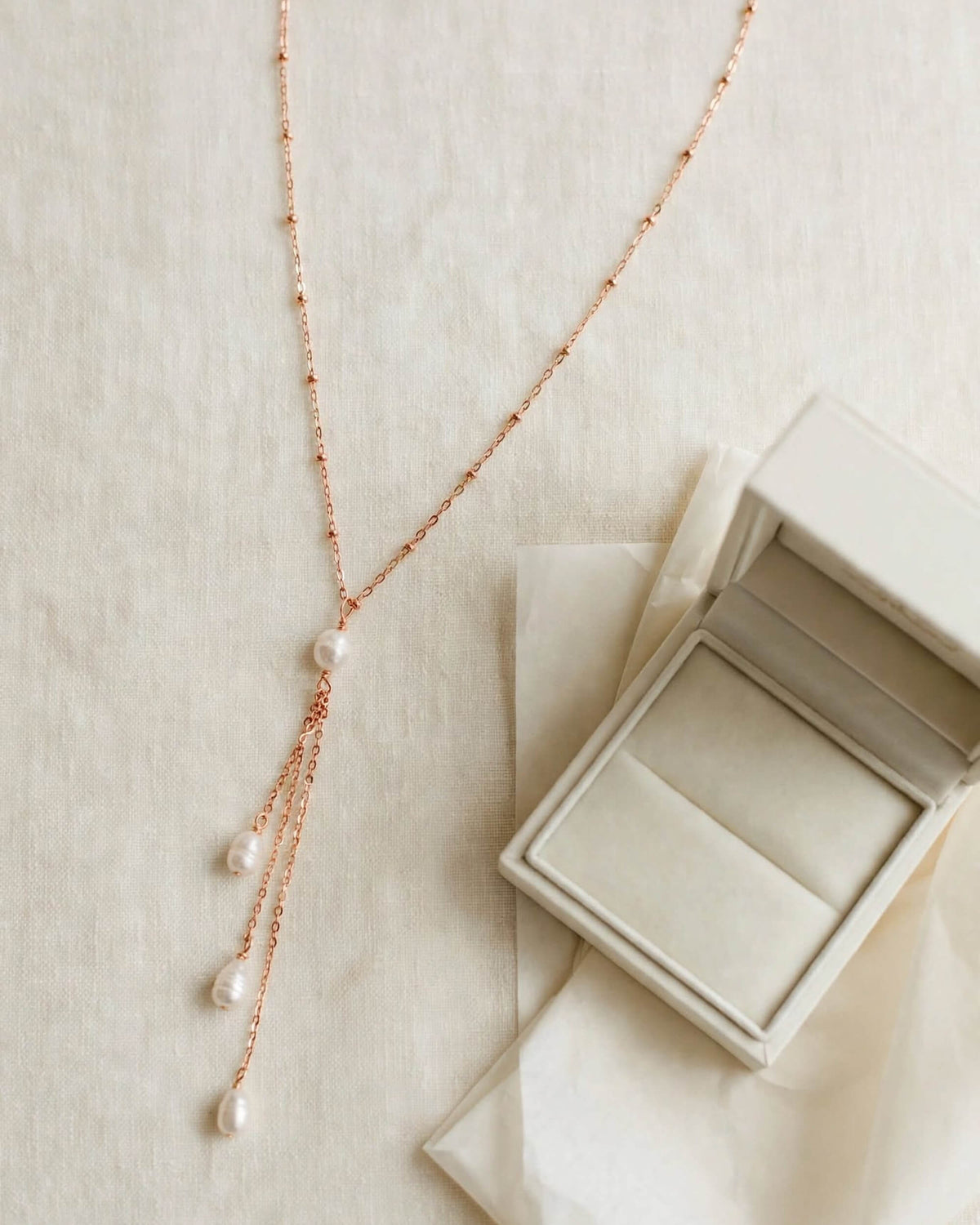 A delicate rose gold lariat necklace with three dangling pearl strands, displayed next to an open cream-colored jewelry box on a textured beige fabric background.