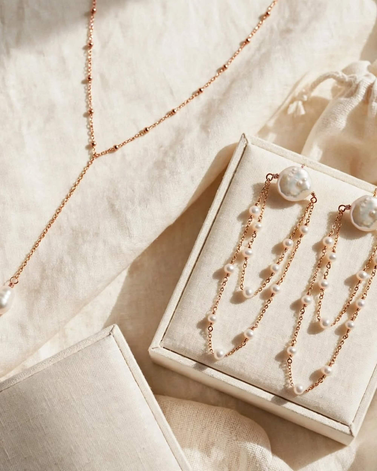 Rose gold jewelry set with pearl accents, including a delicate necklace and matching drop earrings, displayed on a cream-colored jewelry box over a soft linen fabric background.