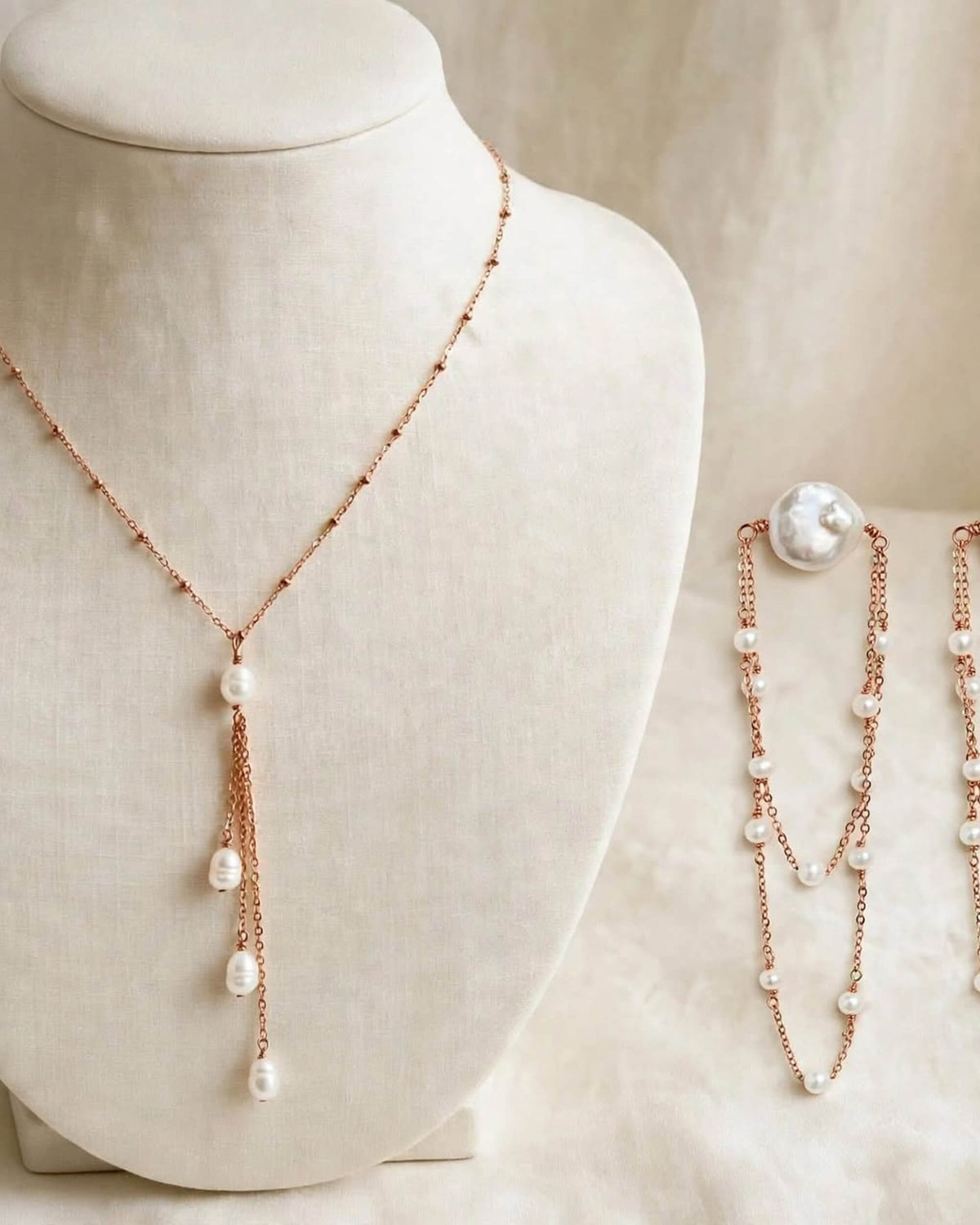 Rose gold double-strand necklace with white baroque pearls, displayed on a cream fabric jewelry bust with matching pearl and chain accessories on a soft beige background.