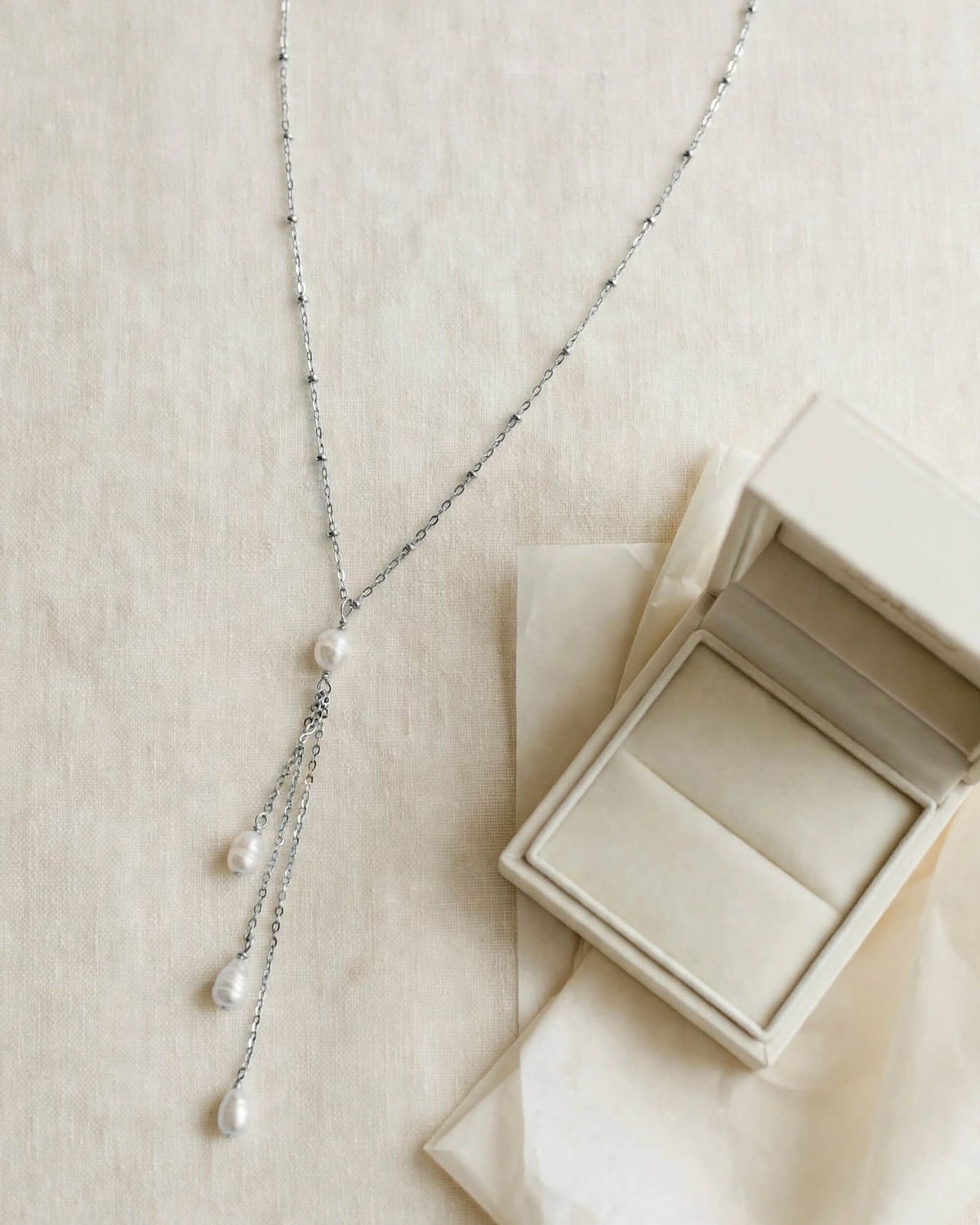 A delicate silver Y-necklace with white pearl drops, displayed next to an open cream-colored jewelry box on a textured beige fabric background.