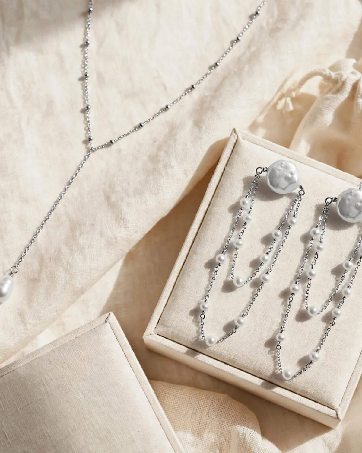 Silver-toned jewelry set including a delicate chain necklace and matching drop earrings with round pearl-like studs and small bead chains, displayed in a cream jewelry box on beige fabric.