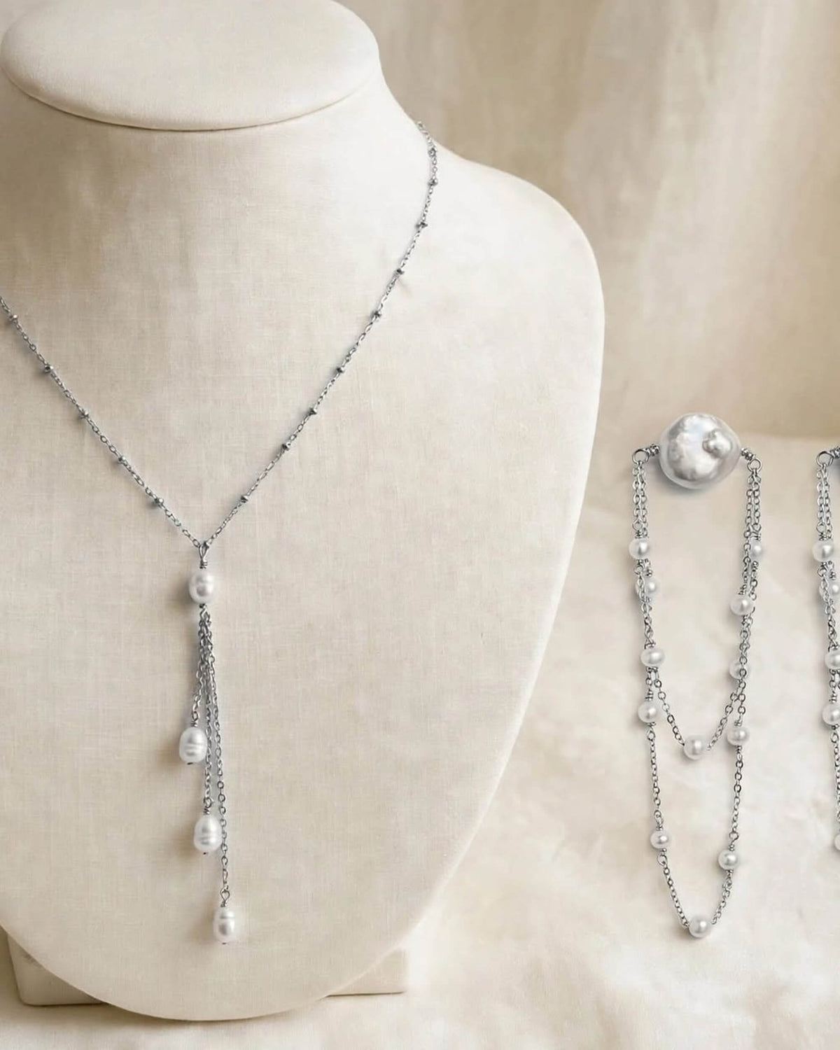 A silver-toned double-strand necklace with white pearl drops, displayed on a cream fabric mannequin bust with a matching close-up inset of the necklace on the right.