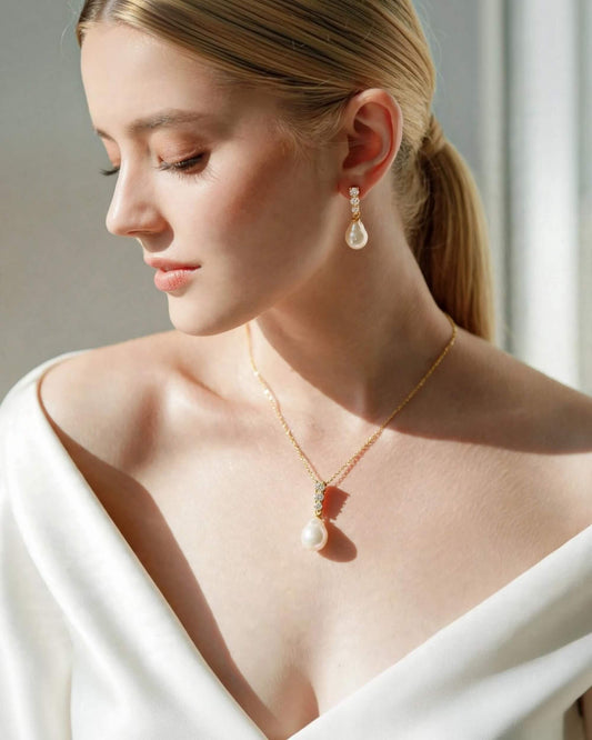 A woman with blonde hair in a ponytail models a gold necklace and matching drop earring with white pearl-like beads, wearing a white off-the-shoulder top in a softly lit indoor setting.