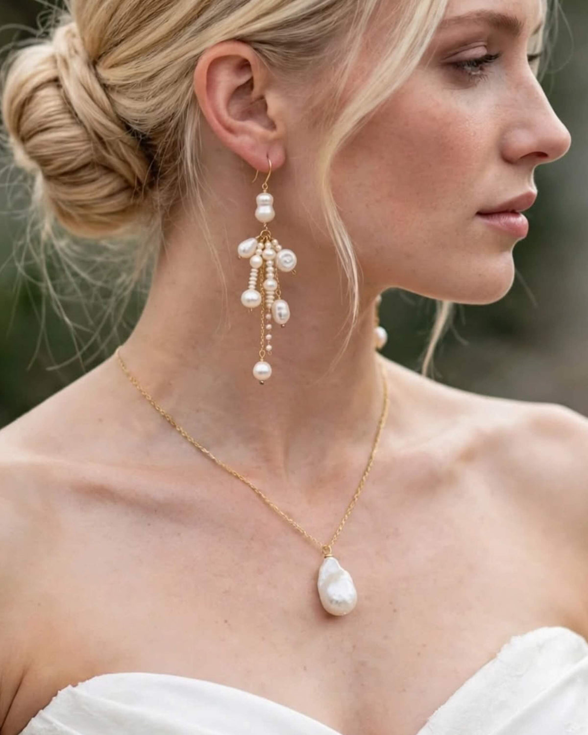A close-up of a woman wearing a gold necklace with a large white baroque pearl pendant and matching dangling pearl and gold earrings.