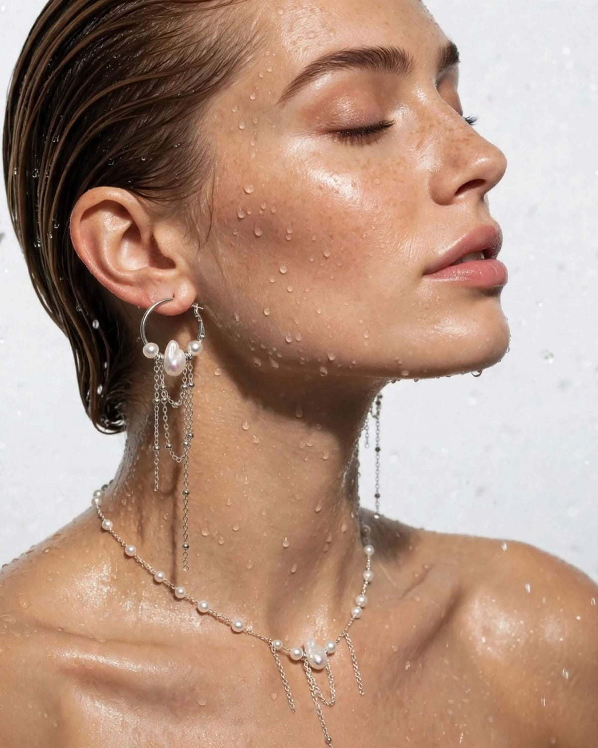 A close-up of a woman with wet hair and water droplets on her skin, wearing a silver-toned drop earring and matching pearl-and-chain necklace against a light gray background.