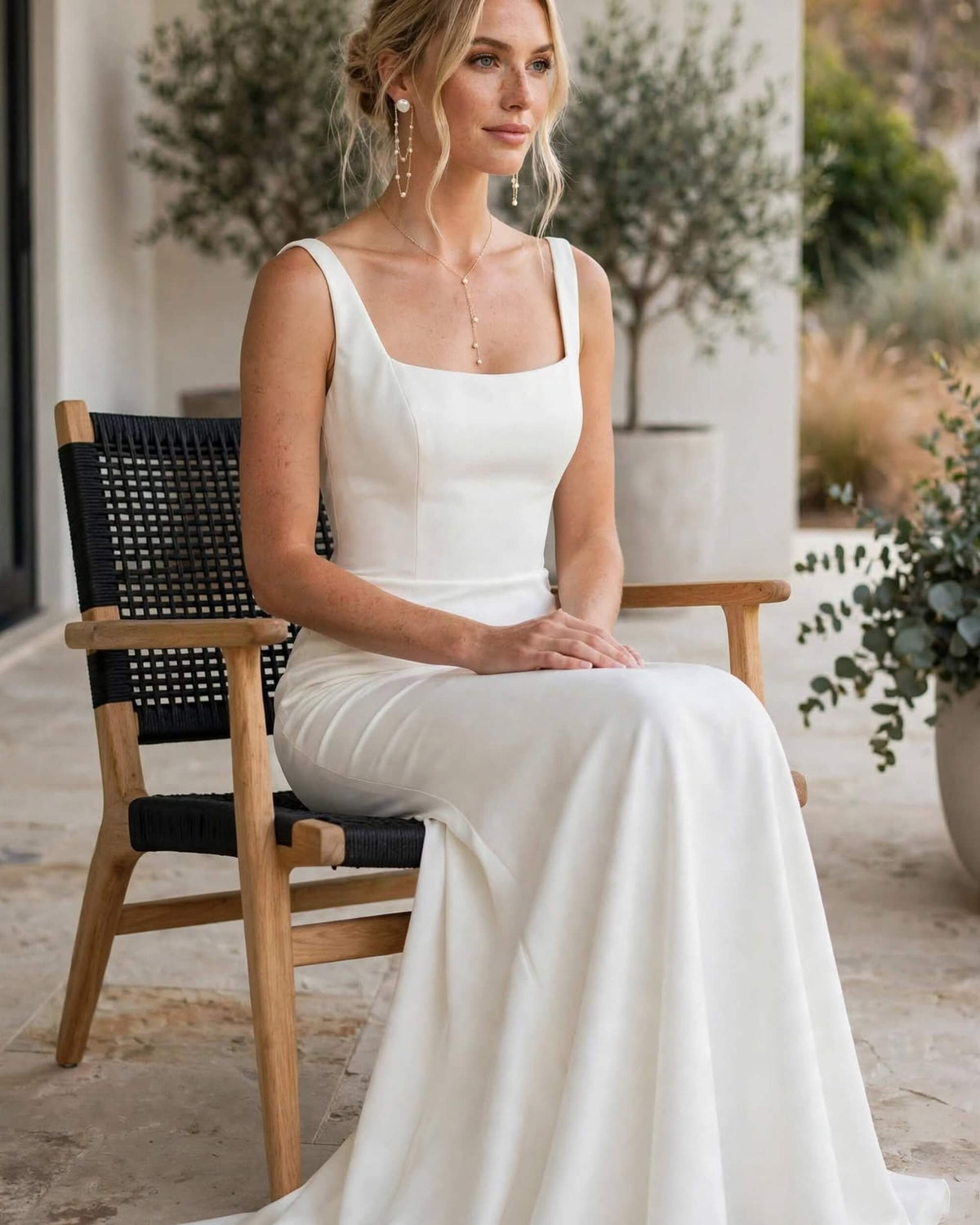 A woman wearing a sleeveless white wedding dress sits on a wooden chair with black woven seats in an outdoor patio setting.