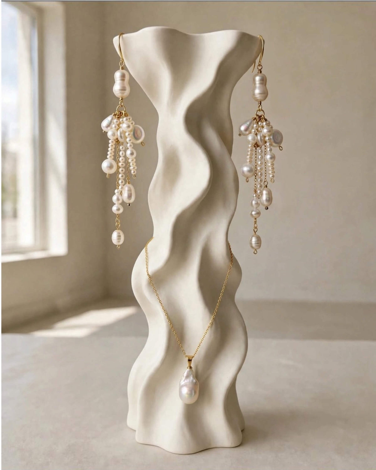 A jewelry display stand shaped like a wavy vase, showcasing a gold necklace with a large pearl pendant and matching pearl drop earrings with gold chains, set against a softly lit beige background.