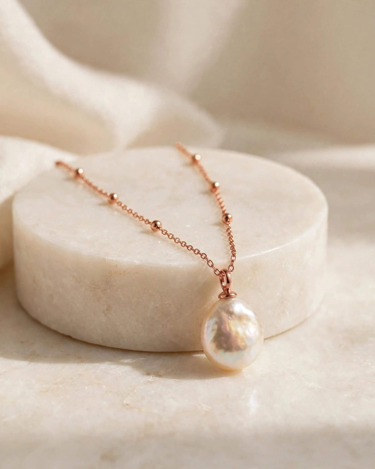 A delicate rose gold chain necklace with a large, irregular lustrous pearl pendant, displayed on a round white marble pedestal.