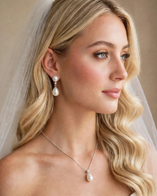 A close-up portrait of a blonde woman wearing a bridal jewelry set with a pearl drop earring and matching pearl pendant necklace on a delicate silver chain, with a sheer veil draped over her head against a soft beige background.