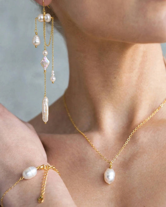 A close-up lifestyle shot of a model wearing a gold jewelry set featuring a dangle earring, a necklace with a baroque pearl pendant, and a matching bracelet on the wrist.