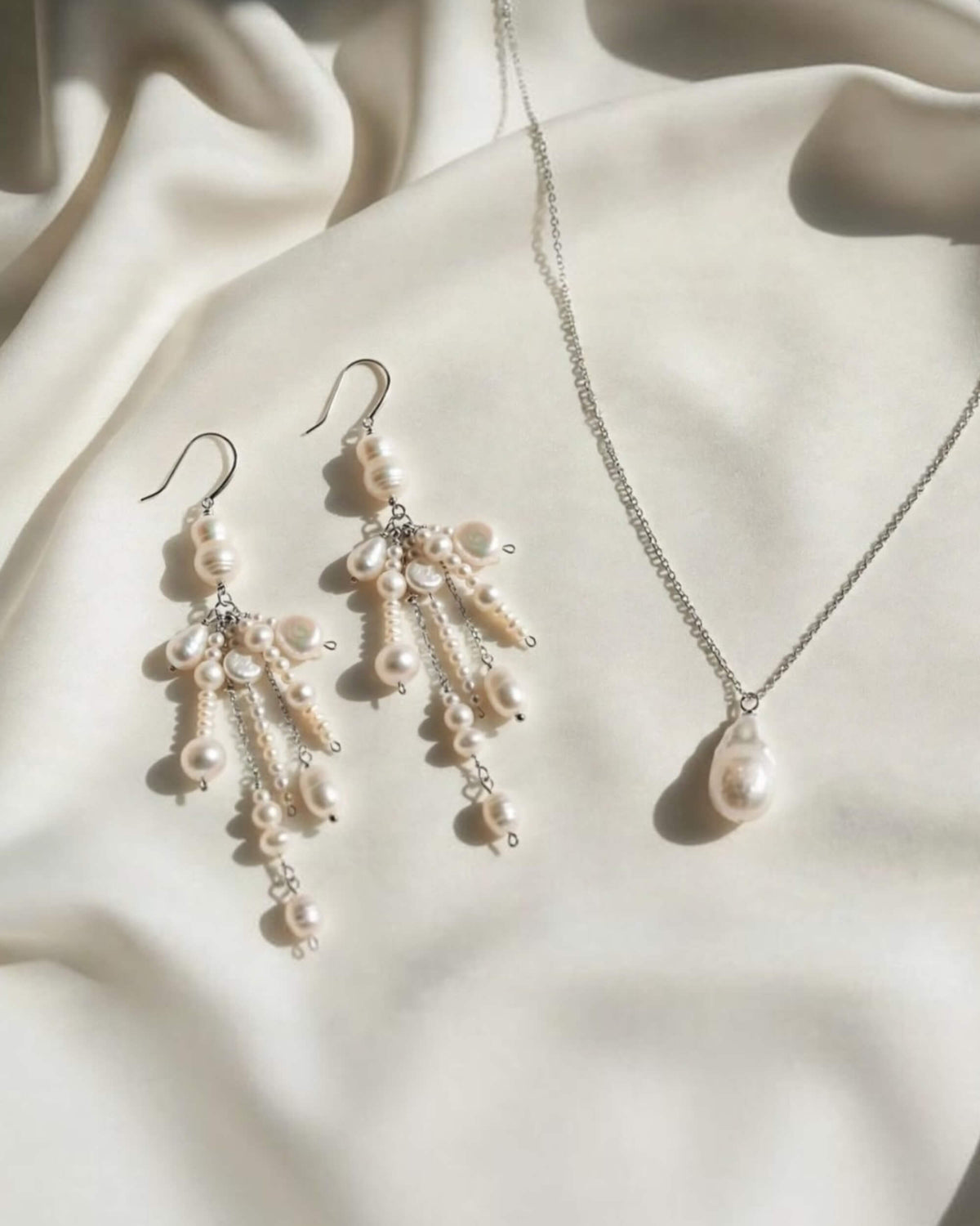 A jewelry set featuring a silver chain necklace and matching drop earrings, both adorned with cream-colored freshwater pearls and small silver beads, displayed on draped cream satin fabric.
