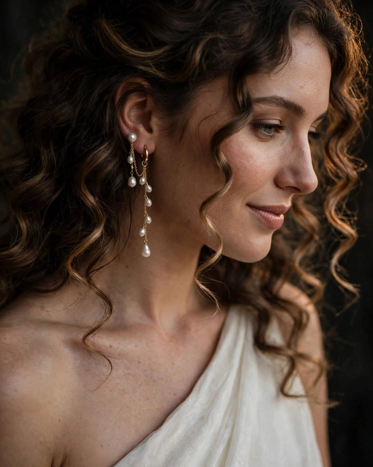 A close-up portrait of a woman with curly brown hair wearing a long pearl drop earring with gold-toned hooks, against a dark background.