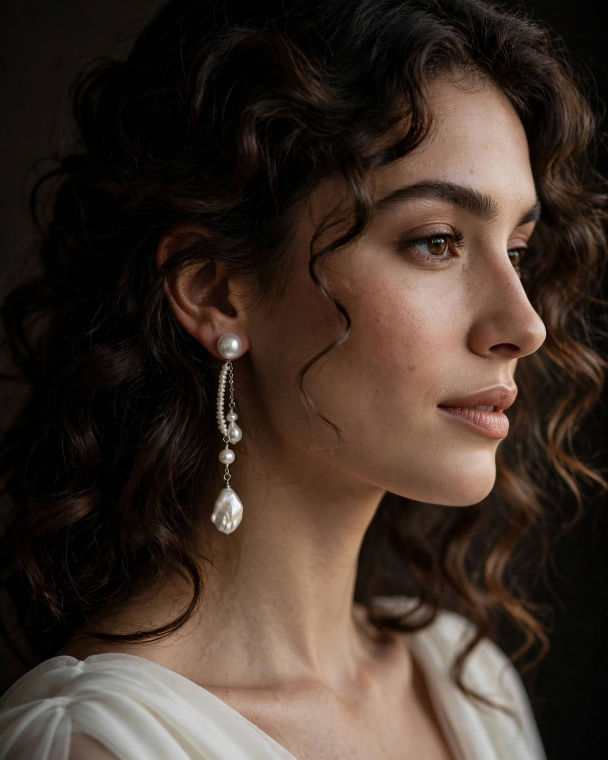 A close-up portrait of a woman with dark curly hair wearing a long pearl drop earring with silver-toned chain details, set against a dark background.