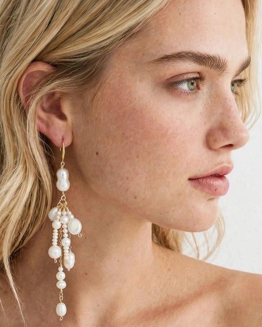 A close-up side profile of a blonde woman wearing a long, dangling pearl earring with gold-tone hooks and beads.