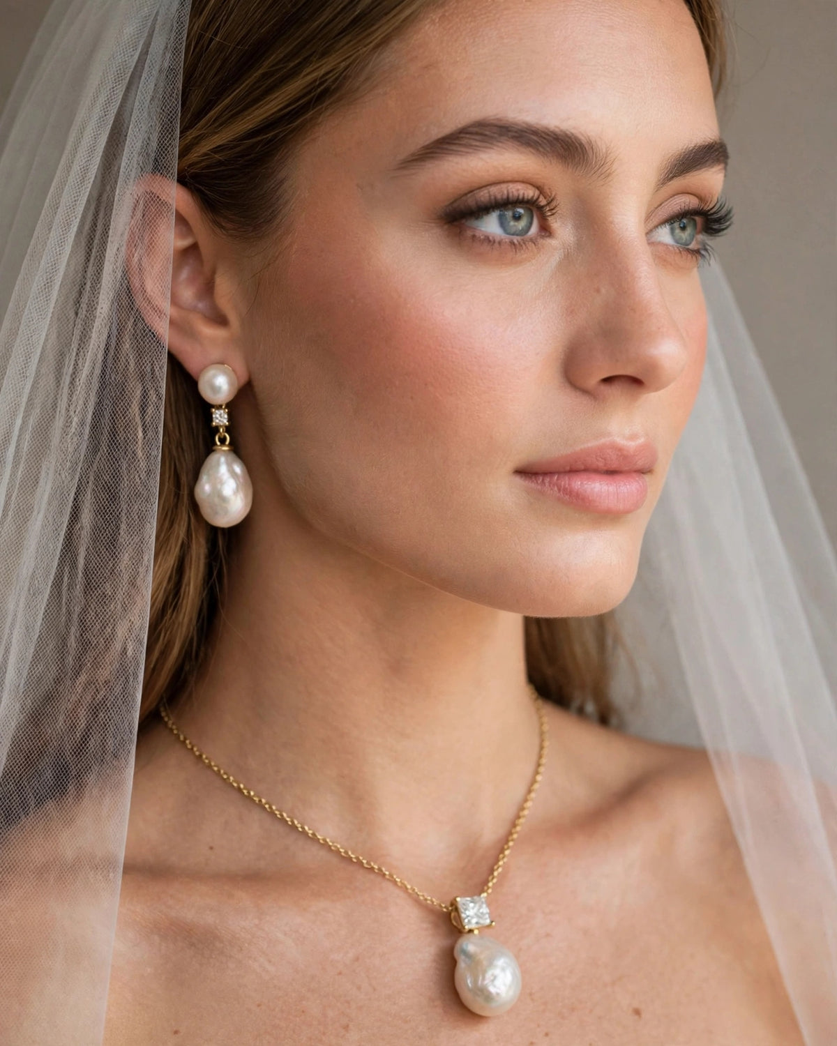 A close-up portrait of a woman wearing a delicate gold necklace with a large baroque pearl pendant and matching drop earrings, along with a sheer white bridal veil.