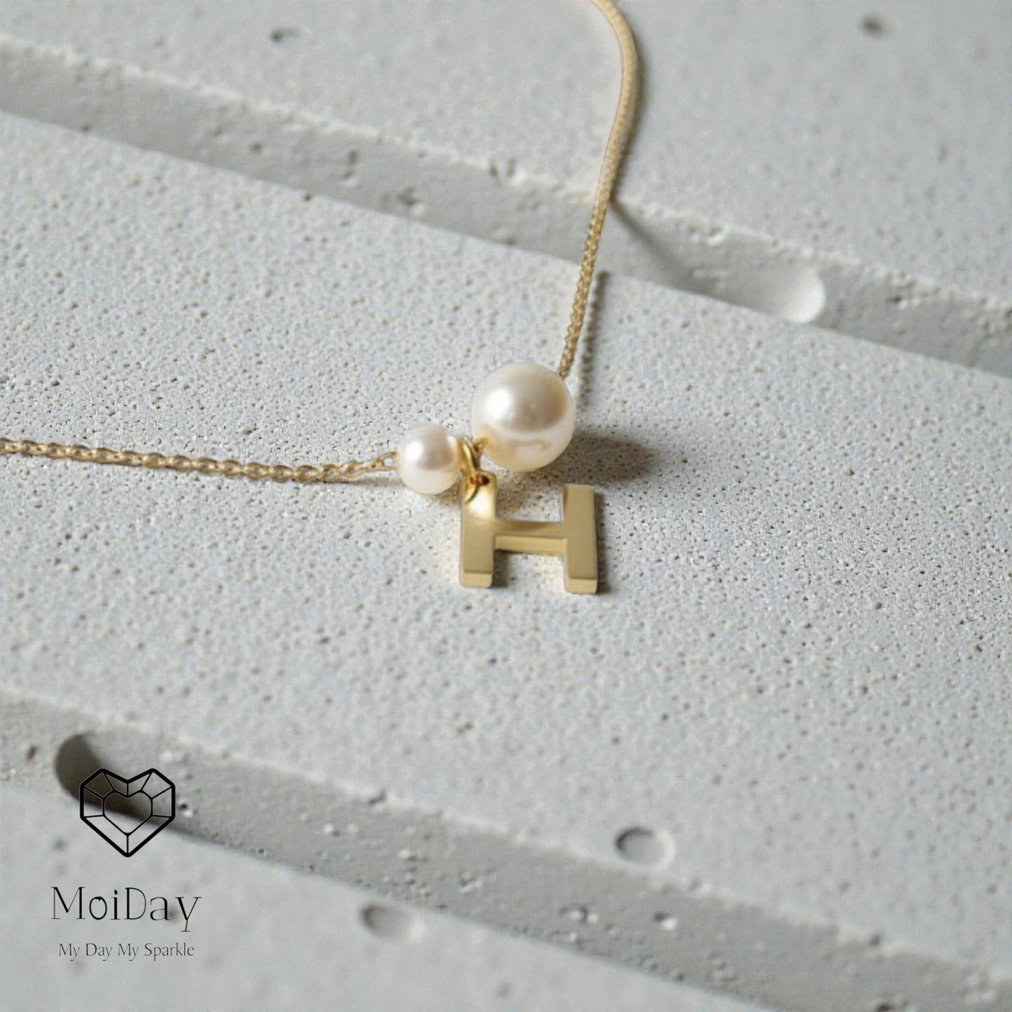 Necklace with two pearls and a gold 'H' pendant on a textured surface, MoiDay brand visible.