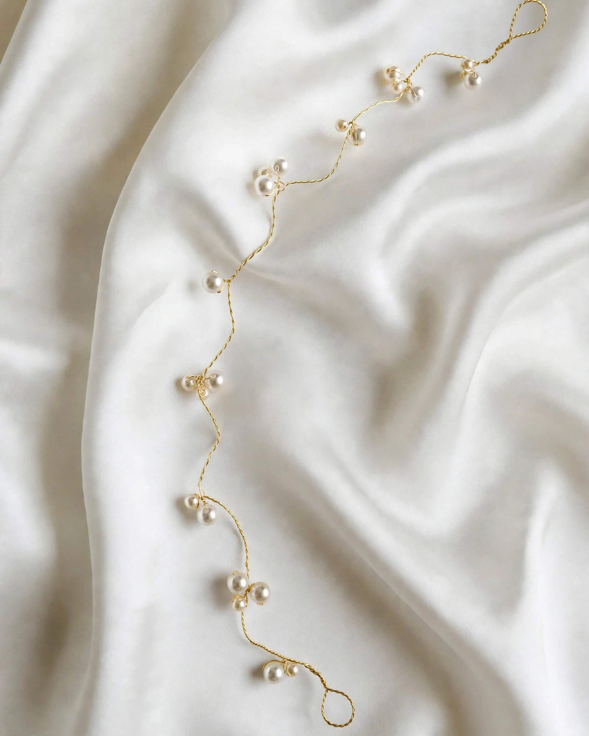 A delicate gold chain headband embellished with small white pearls, laid out diagonally on a draped white satin fabric background.