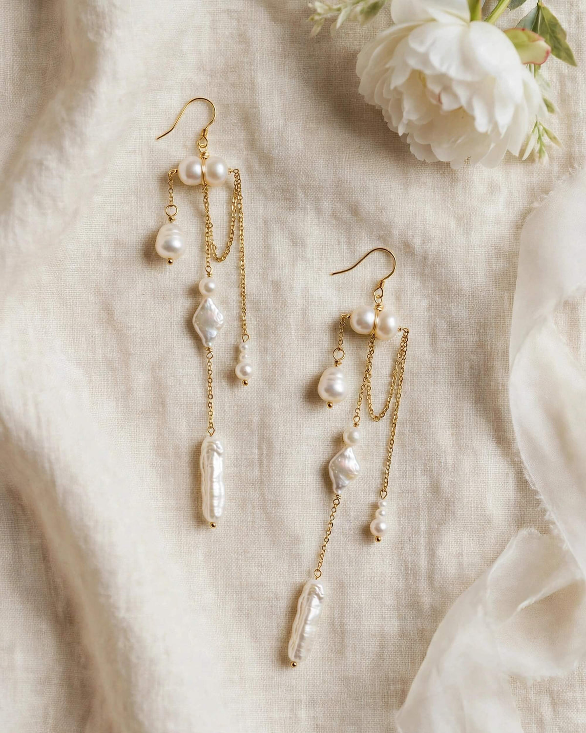 Gold dangle earrings with baroque pearls and clear crystal drops, displayed on a textured beige linen fabric next to a white flower.