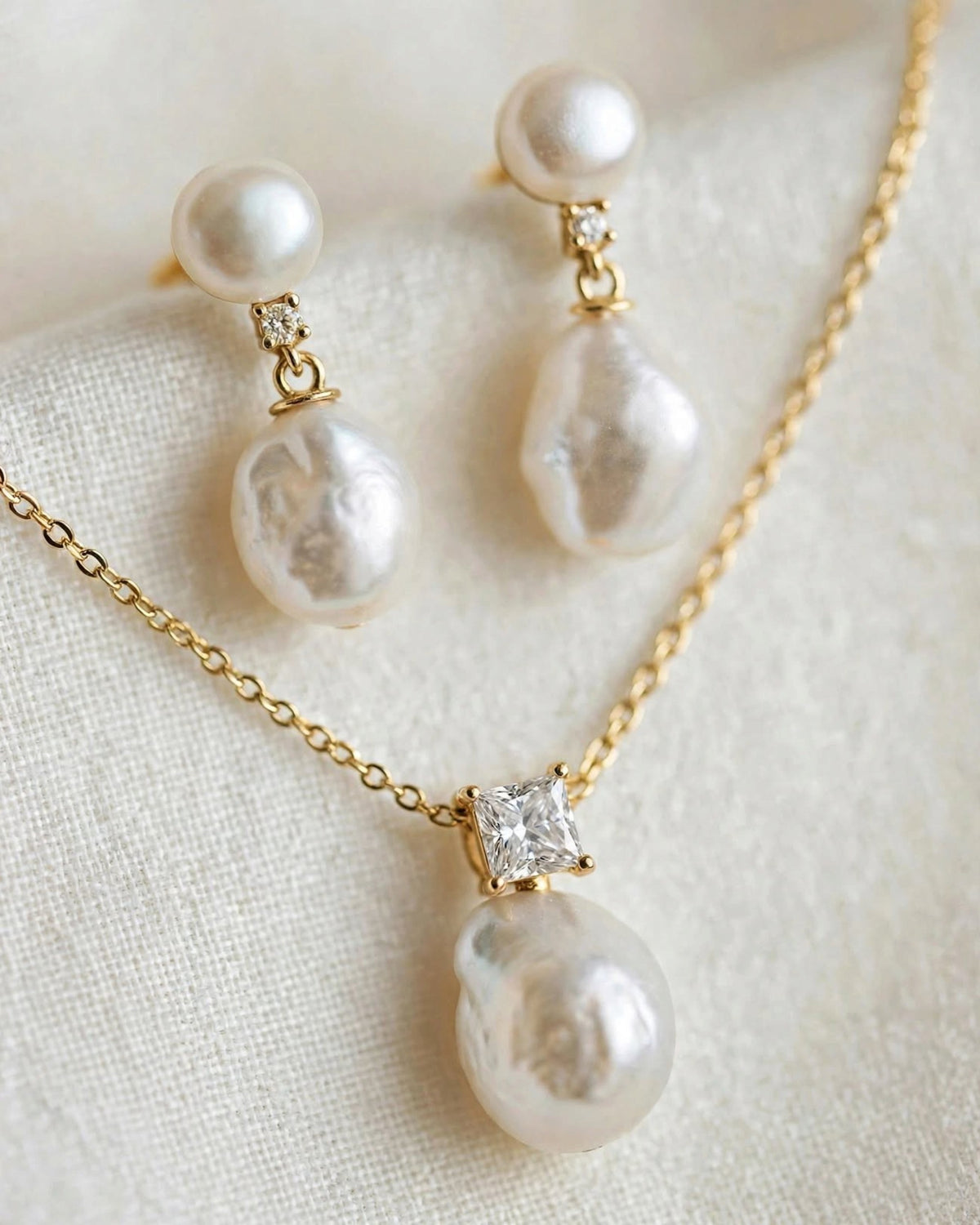 Gold-tone jewelry set featuring a necklace and matching earrings with large baroque-style white pearls and small clear accent stones, displayed on a soft cream fabric background.