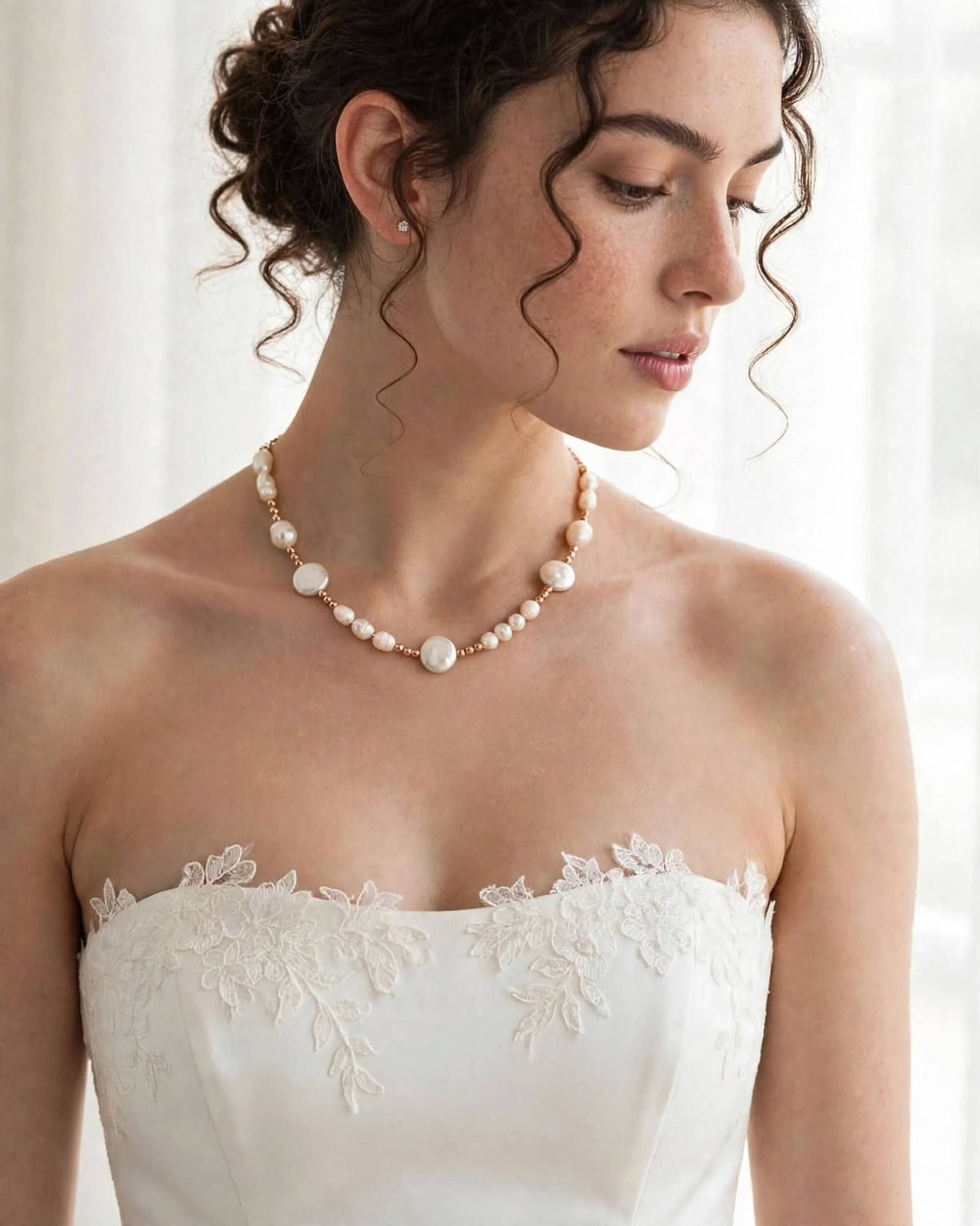 A woman wearing a pearl and gold statement necklace with a strapless white lace wedding dress, photographed in a bright, softly lit indoor setting.