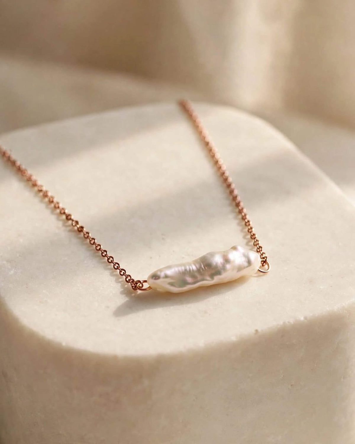 A delicate rose gold chain necklace featuring a natural, irregular-shaped white baroque pearl pendant, displayed on a textured cream-colored stone block.