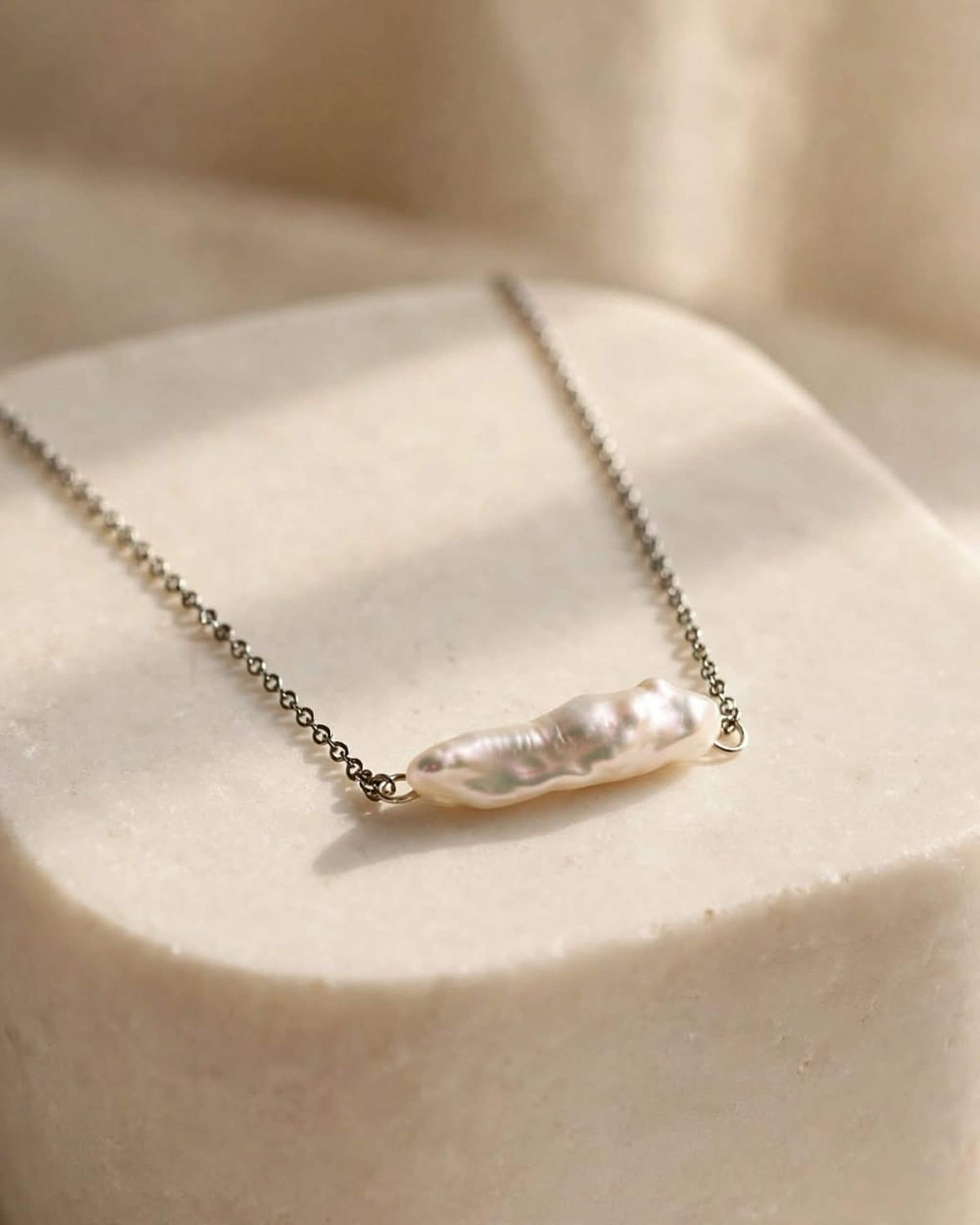 A delicate silver chain necklace featuring a polished, irregular baroque pearl pendant, displayed on a textured beige stone pedestal.
