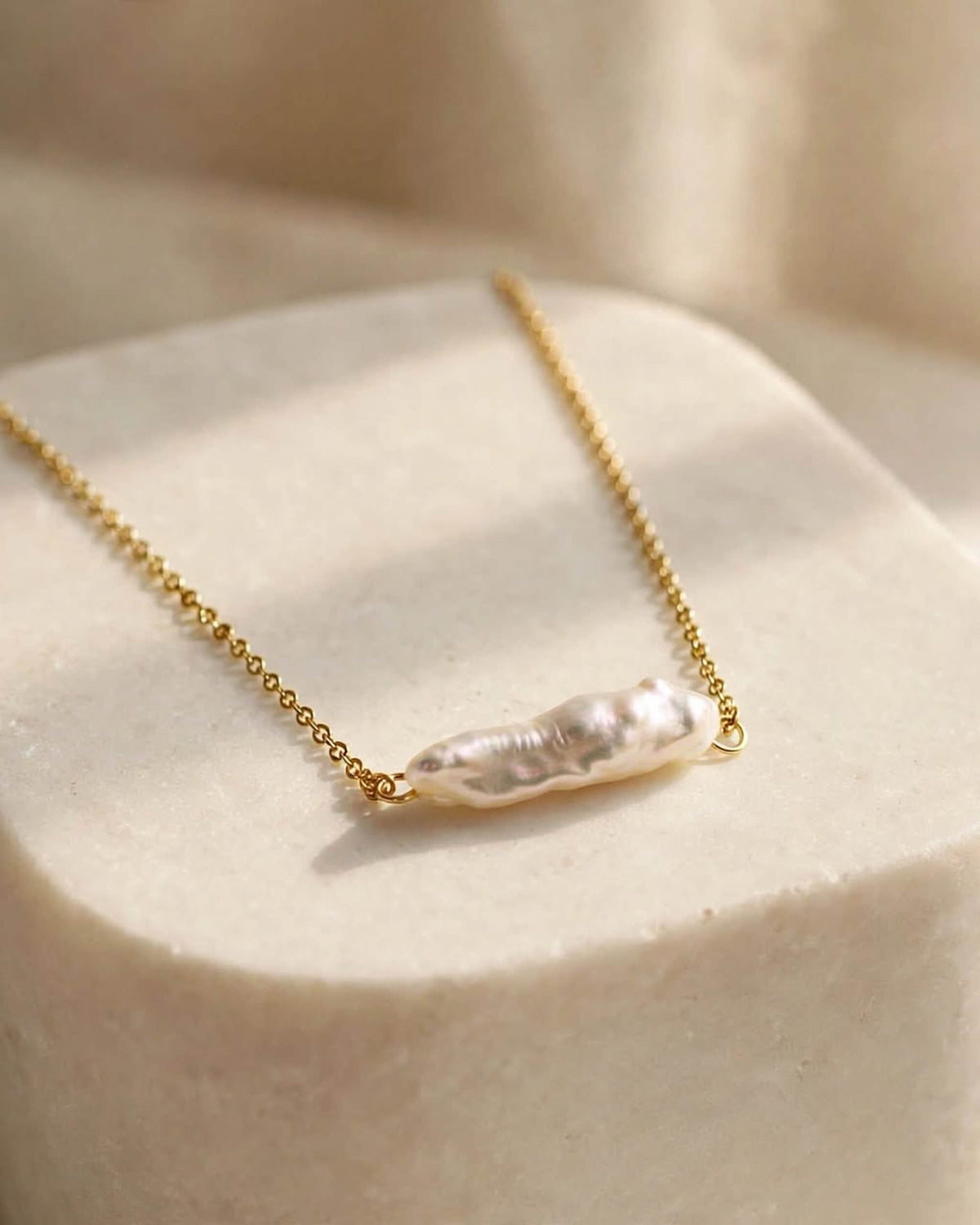 A delicate gold chain necklace featuring a natural, irregular-shaped white baroque pearl pendant, displayed on a textured cream-colored block.