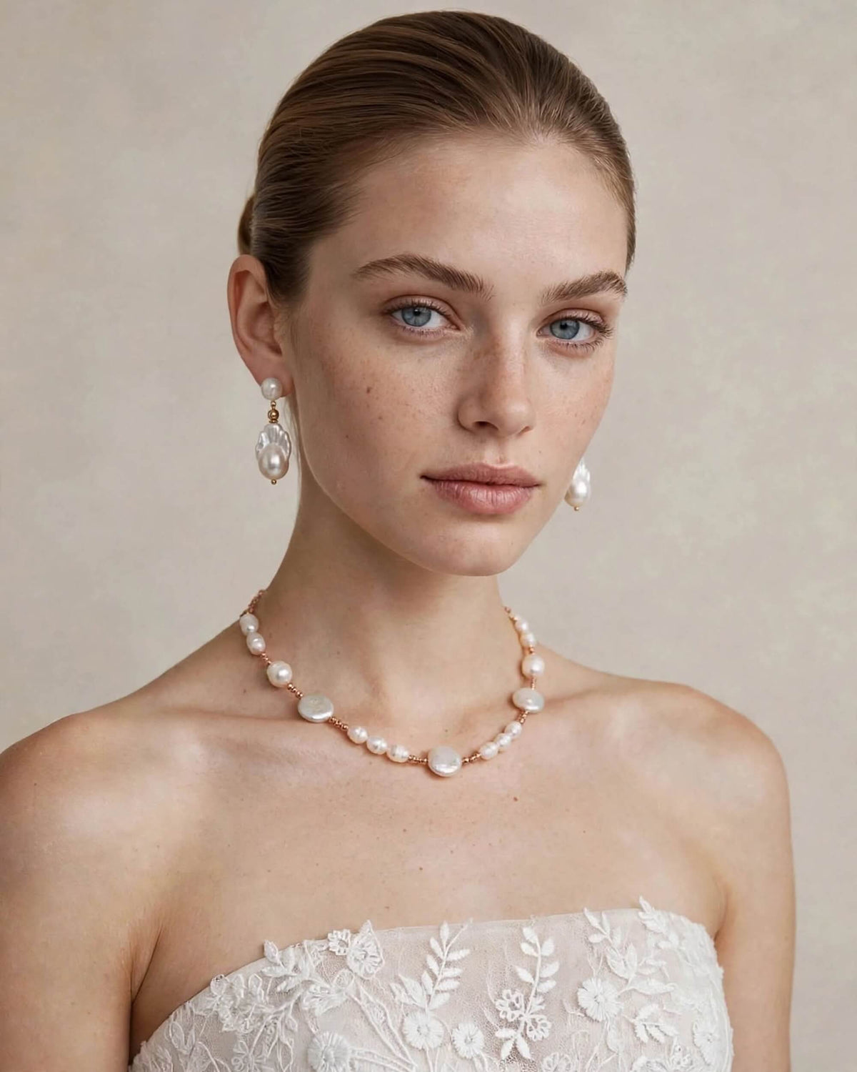 A woman with her hair pulled back wears a pearl necklace and matching pearl drop earrings, with a strapless white lace dress visible at the bottom against a soft beige studio background.