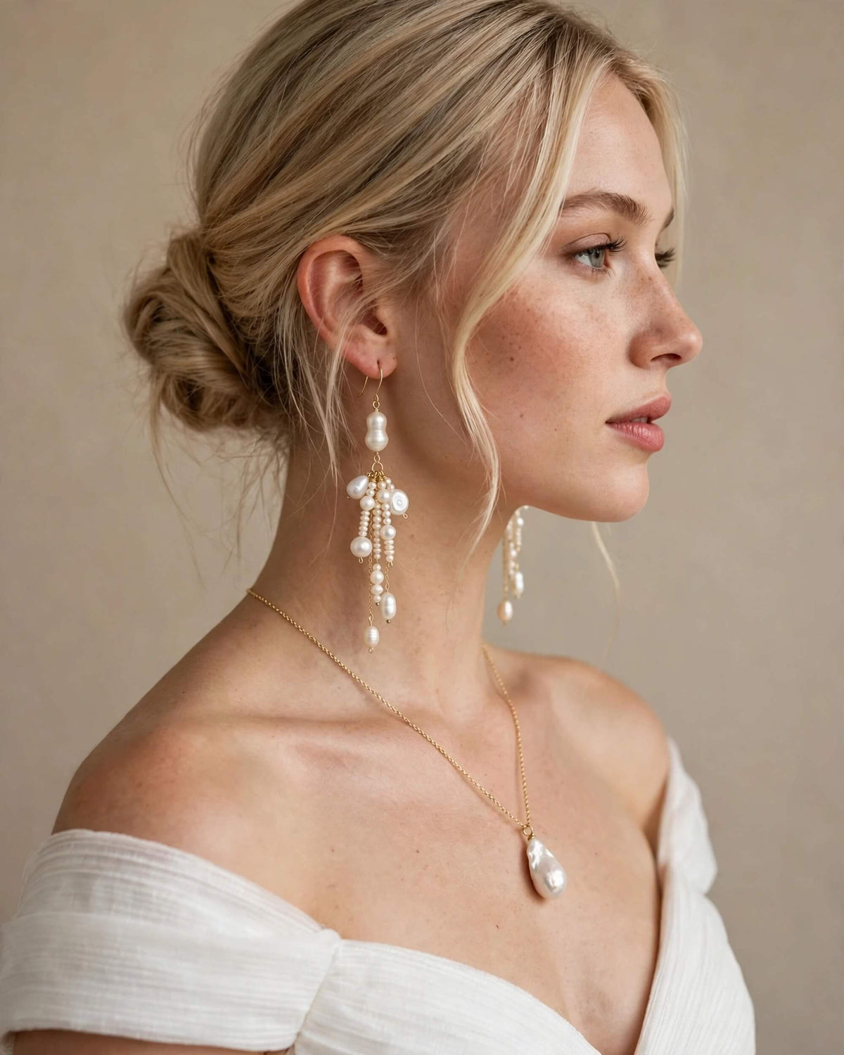 A woman in profile wearing elegant pearl drop earrings and a matching pearl teardrop pendant necklace with a delicate gold chain, styled with a white off-the-shoulder top against a neutral beige background.