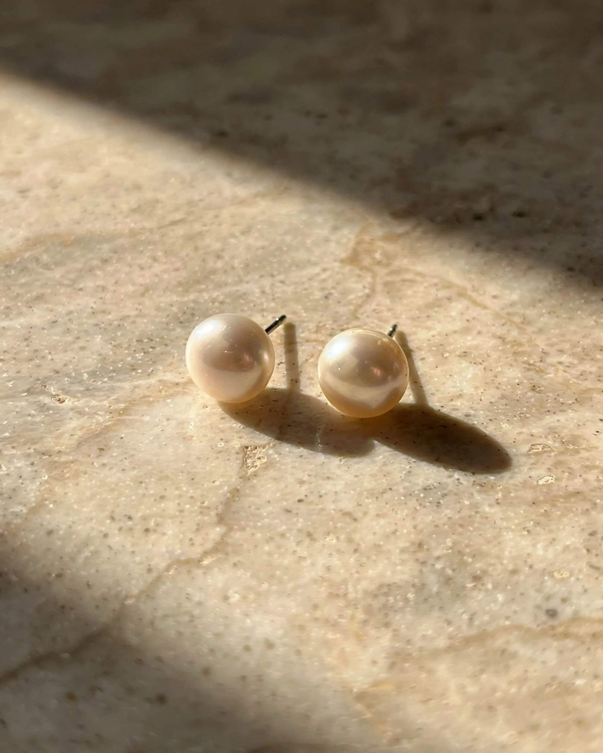 A pair of round freshwater pearl stud earrings with gold posts, resting on a textured beige stone surface under warm, direct sunlight that casts soft shadows.