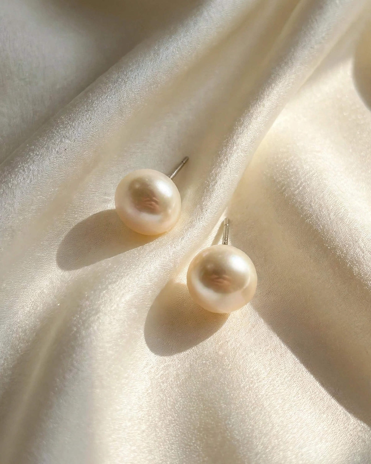 A pair of round freshwater pearl stud earrings with silver posts, resting on draped cream-colored satin fabric.