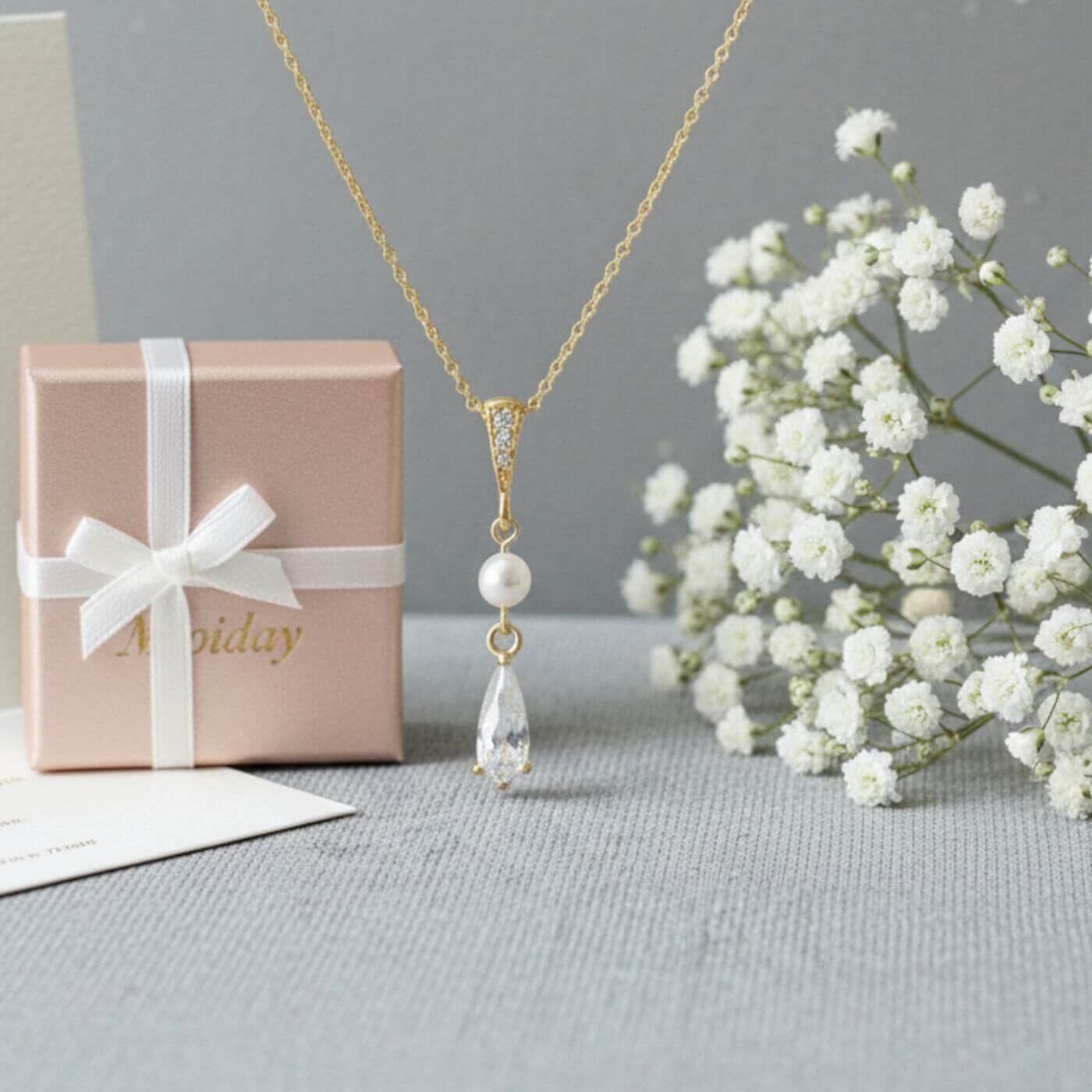 Gold necklace with a pendant, gift box, and white flowers on a gray surface