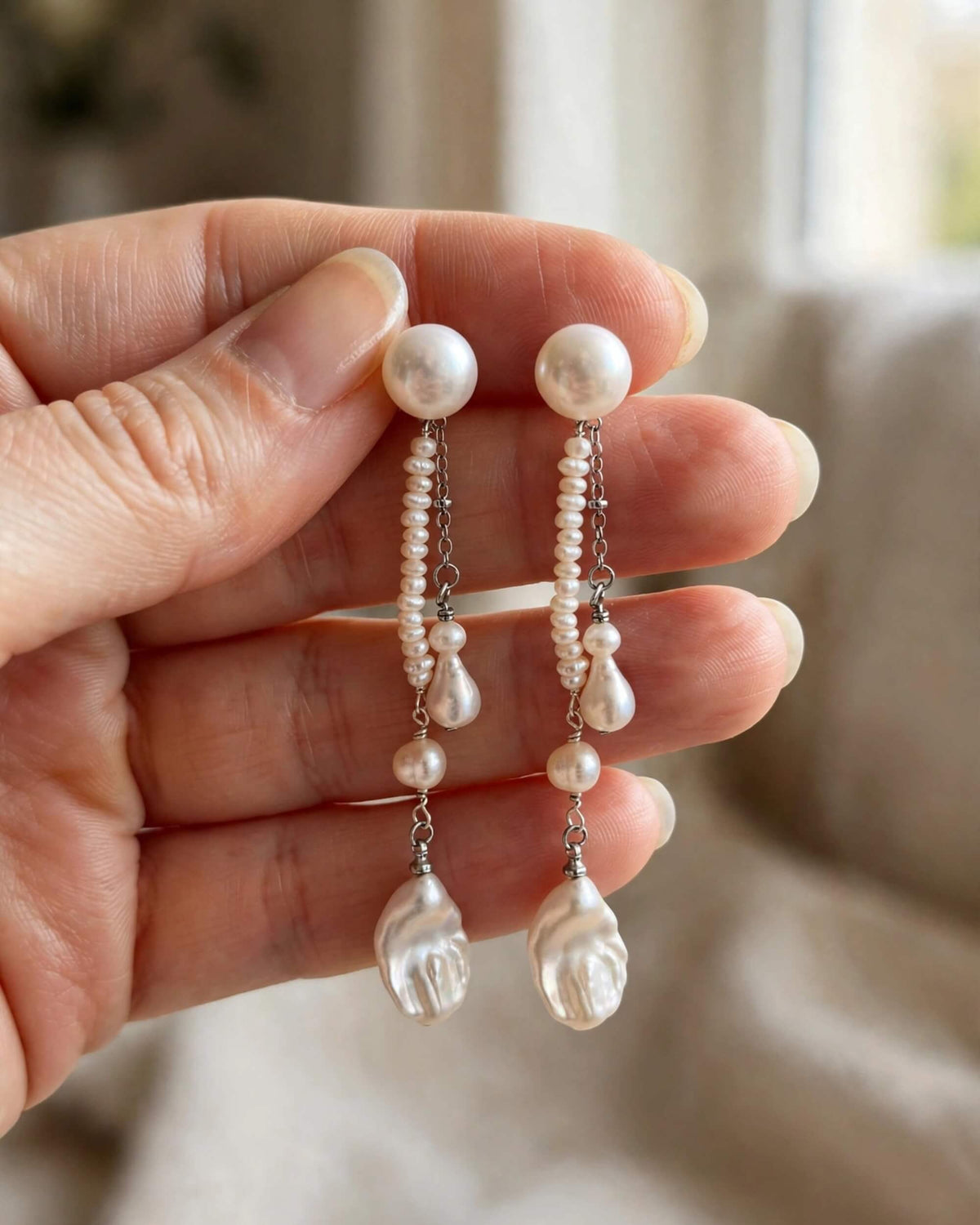 A close-up of a hand holding a pair of dangling pearl earrings with silver-toned chain details and irregular white pearls, against a softly blurred indoor background.