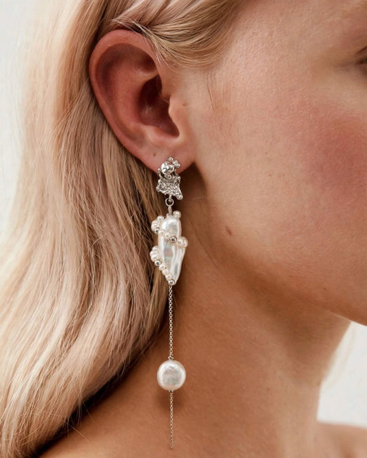 A close-up of a model's ear wearing a long, multi-tiered dangle earring. The earring features a silver-toned textured top, a cluster of white faux pearls, and a long chain ending in a larger white pearl bead.
