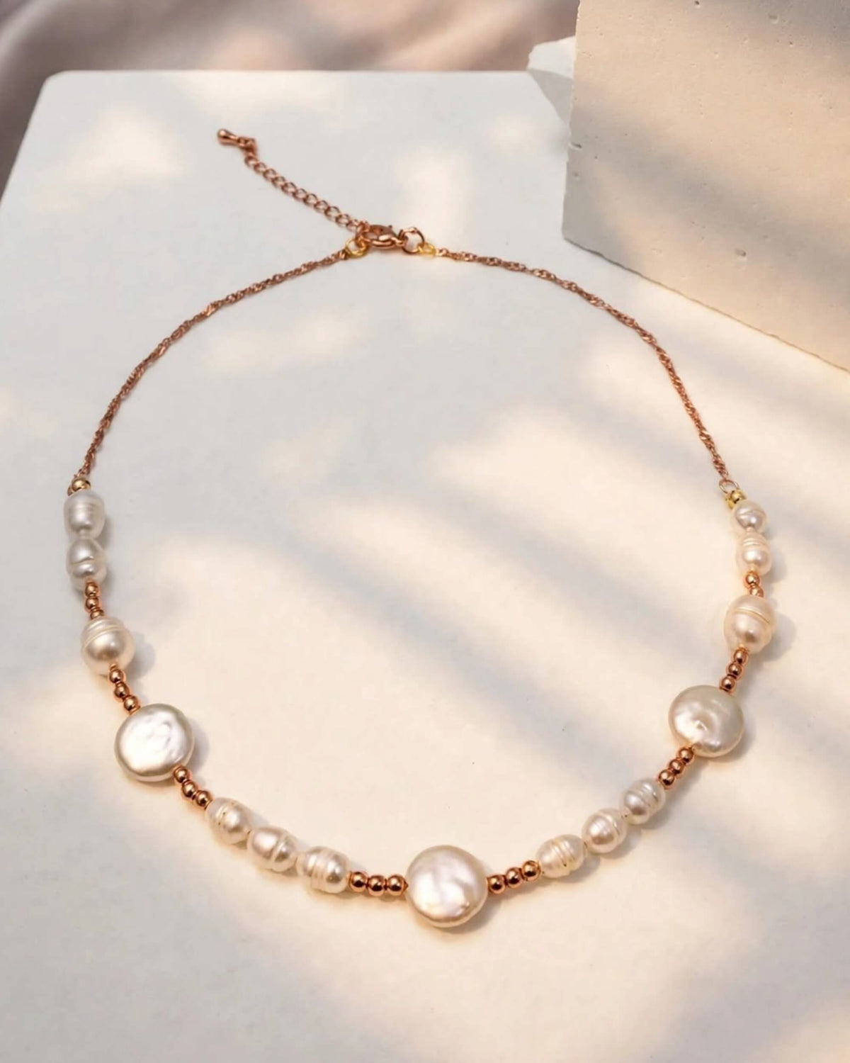 A delicate rose gold chain necklace featuring irregular white baroque pearls and small rose gold beads, displayed on a textured white surface with soft shadows.