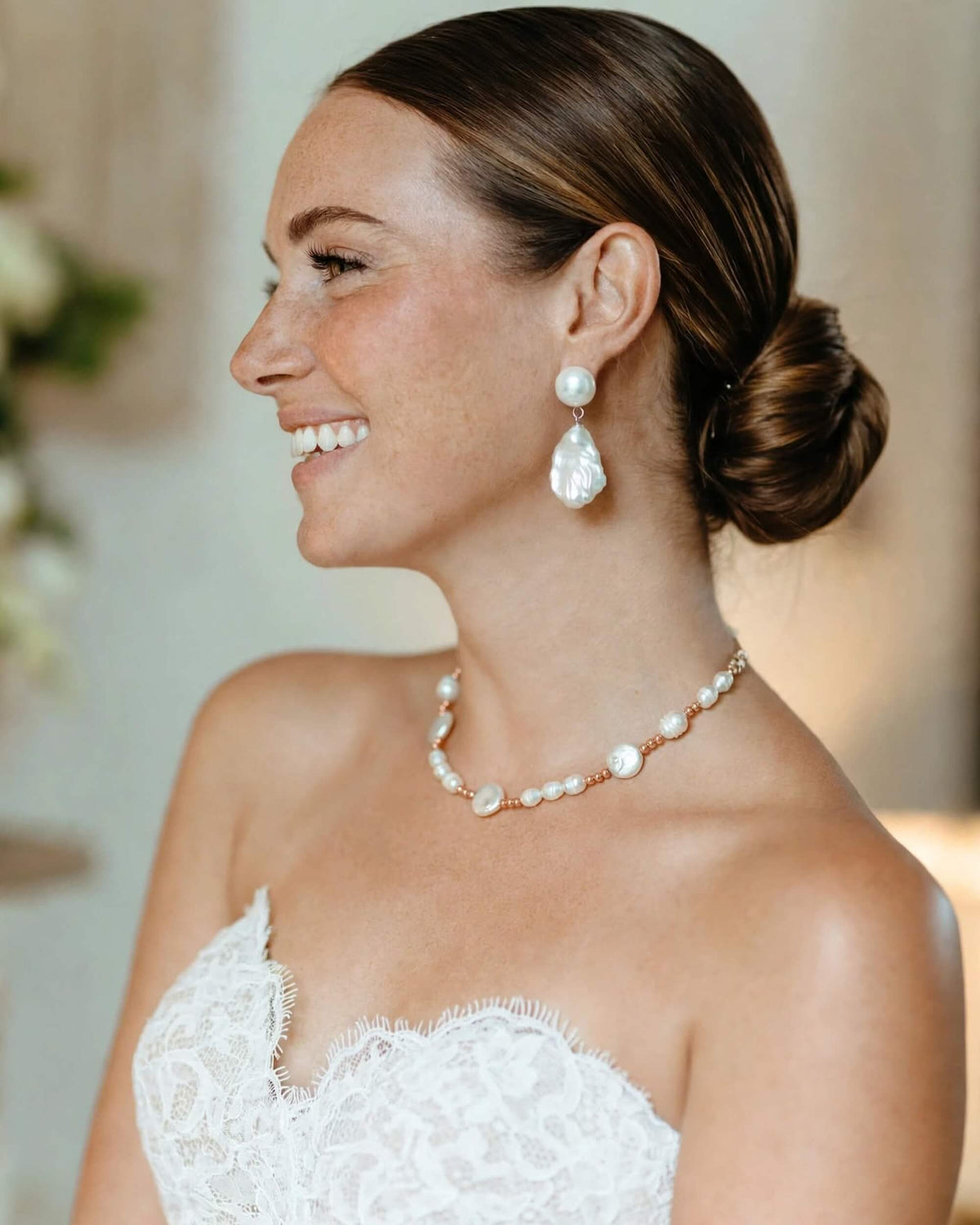 A smiling woman in profile wearing a white lace strapless dress, a pearl necklace, and matching pearl drop earrings with rose gold accents, set against a softly blurred indoor background.