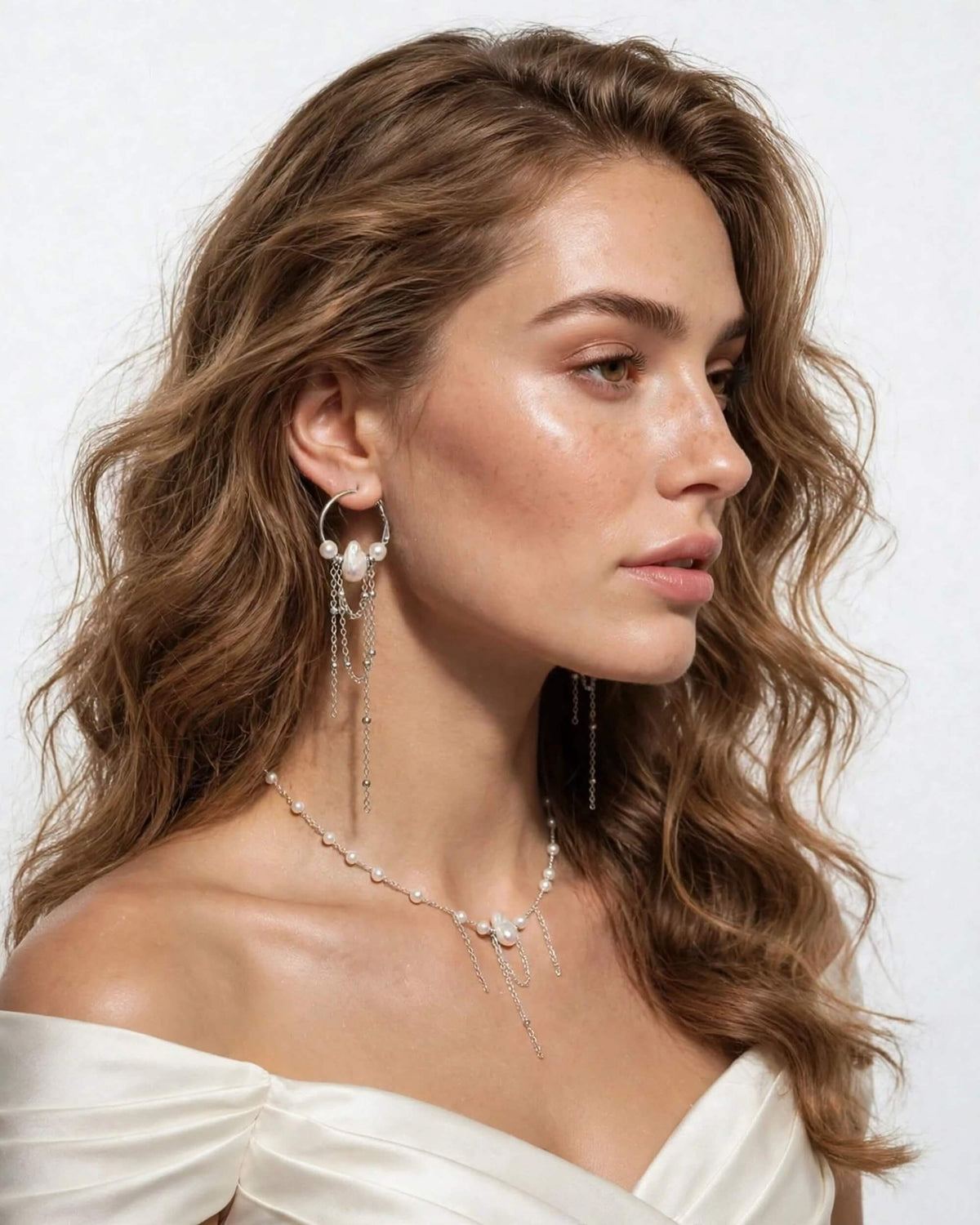 A woman with wavy brown hair is shown in profile wearing a delicate silver-toned necklace and matching drop earrings with pearl-like beads, against a clean white background.