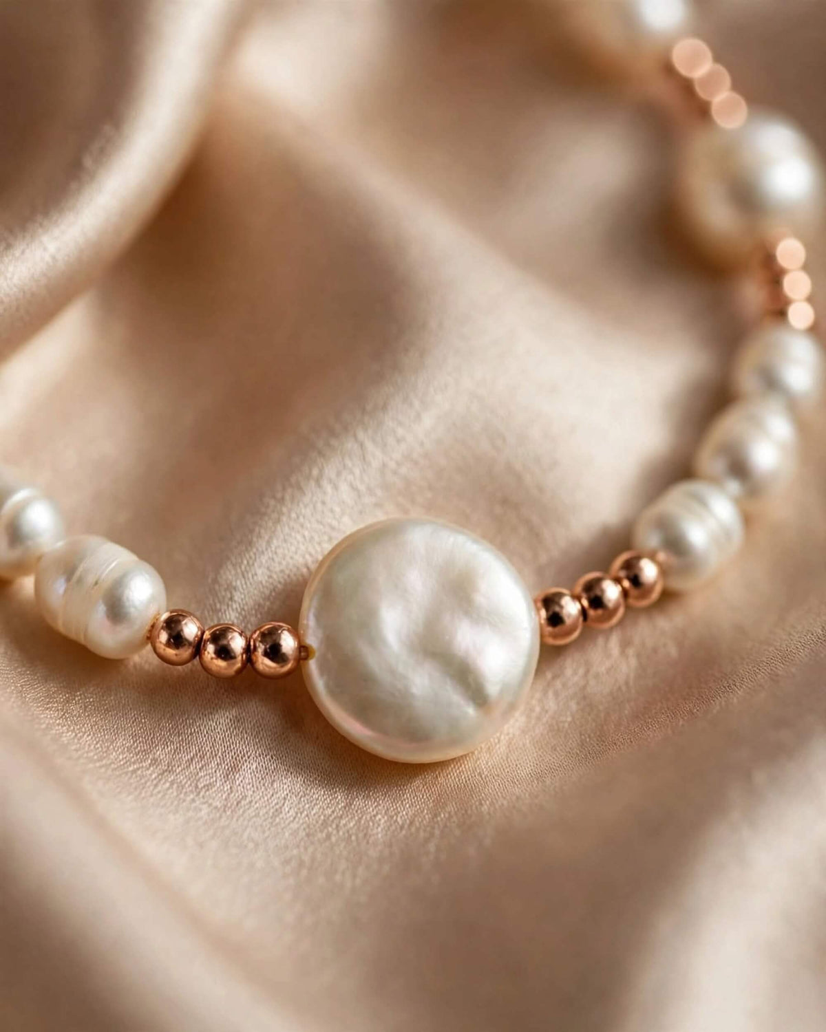 A close-up of a beaded necklace featuring lustrous white baroque pearls and small polished rose gold beads, displayed on draped champagne satin fabric.