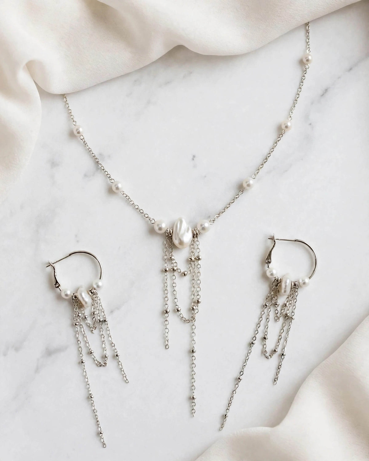 Silver-tone jewelry set featuring a delicate necklace and matching hoop earrings with dangling chain fringe and small pearl accents, arranged on a white marble surface with soft cream fabric.
