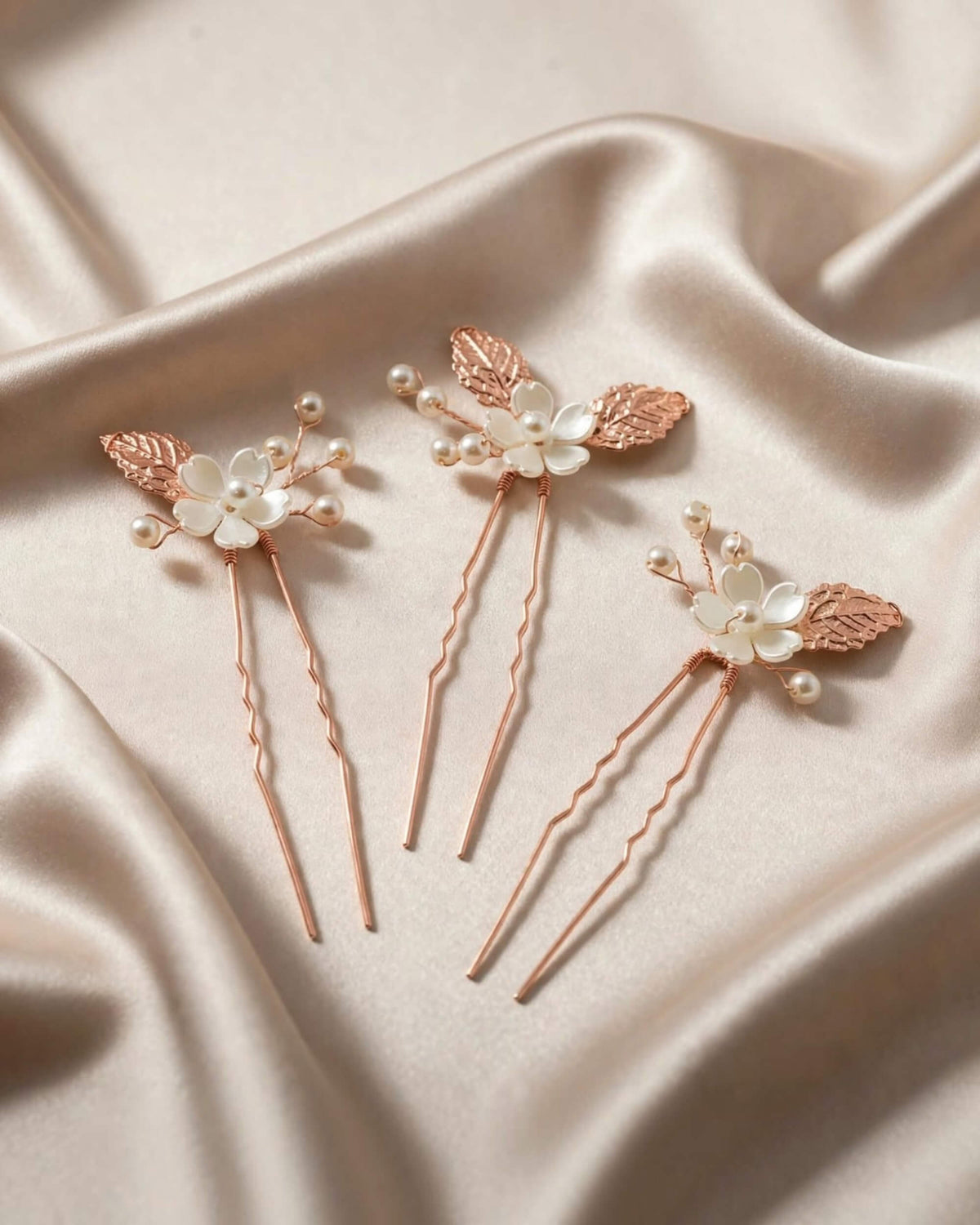 Three rose gold hair pins with white pearl flowers and leaf accents, arranged on a draped beige satin fabric background.